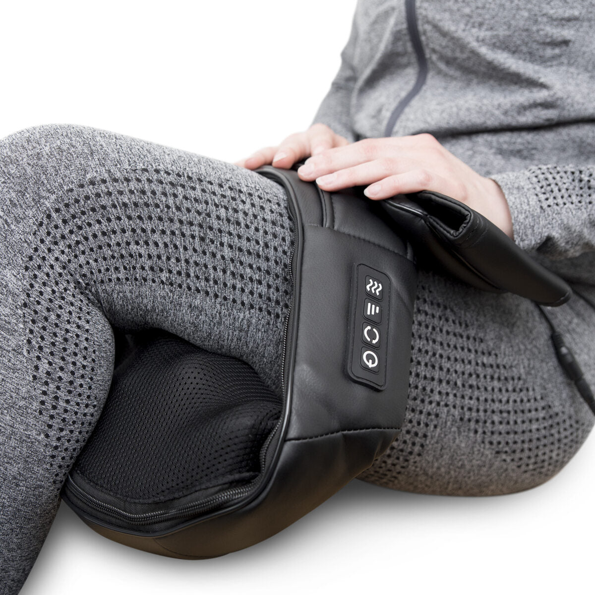 A person in a gray tracksuit uses the Neck & Shoulder Shiatsu Massager with control buttons on their thigh. The black device provides deep kneading muscle relief while their hand rests on it.