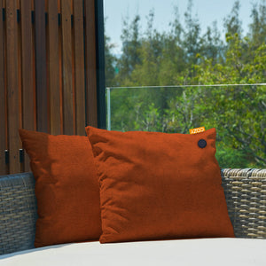 2 orange heated cushions shown on an outdoor rattan sofa with wood panels and trees.