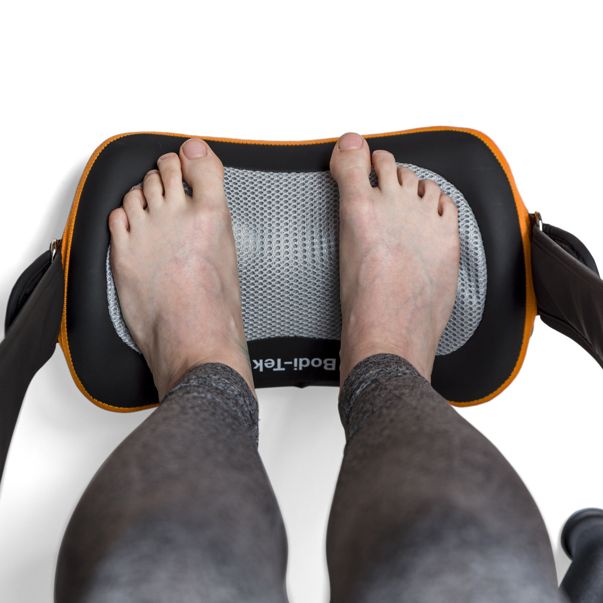 A person in gray leggings rests their bare feet on a black and orange Neck, Shoulder & Body Shiatsu Massager with a mesh cover, viewed from above on a white background.