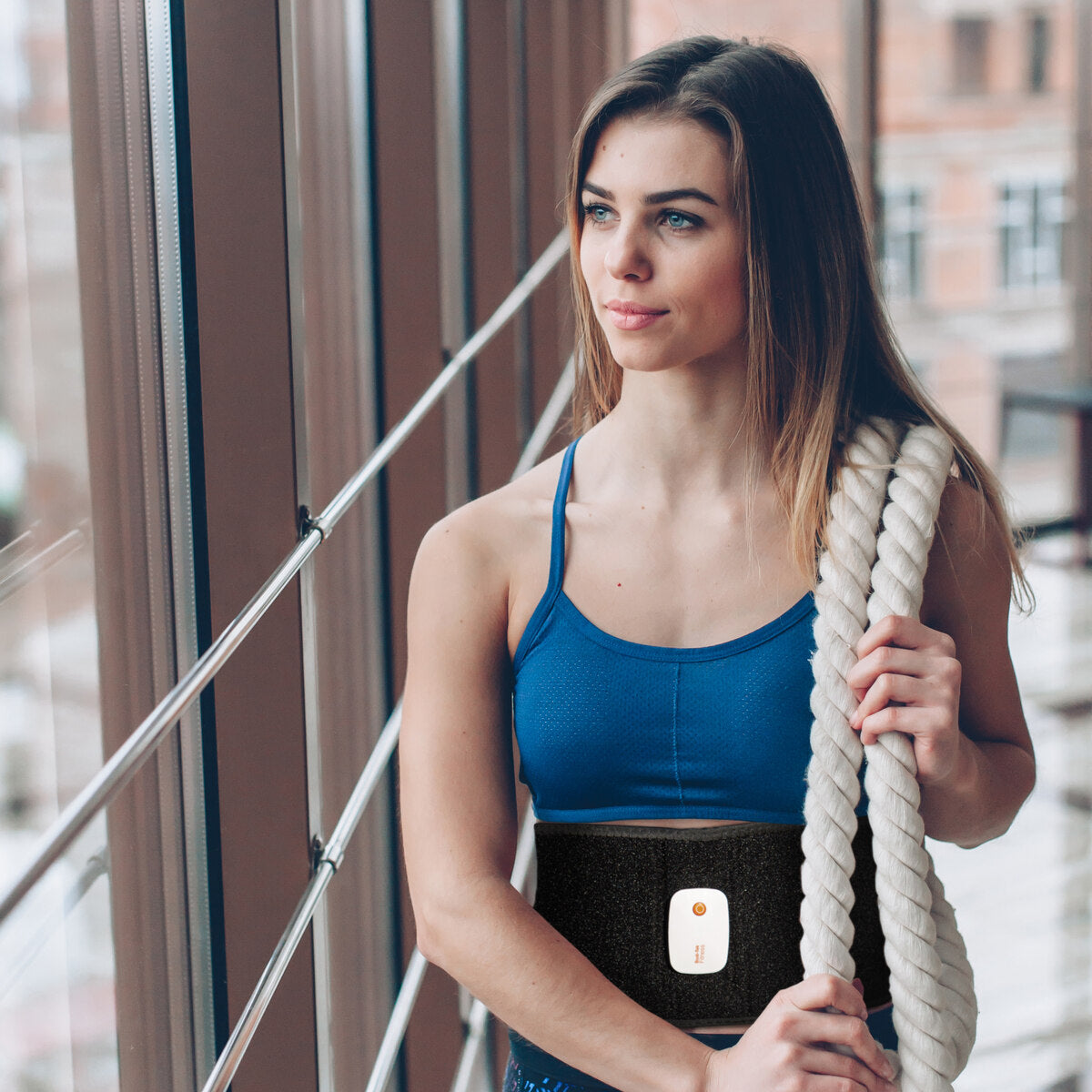 A woman in athletic wear stands indoors by large windows, holding a thick rope and wearing a Smart Fitness EMS Muscle Toning Belt with a small electronic device attached for effective abs firming.