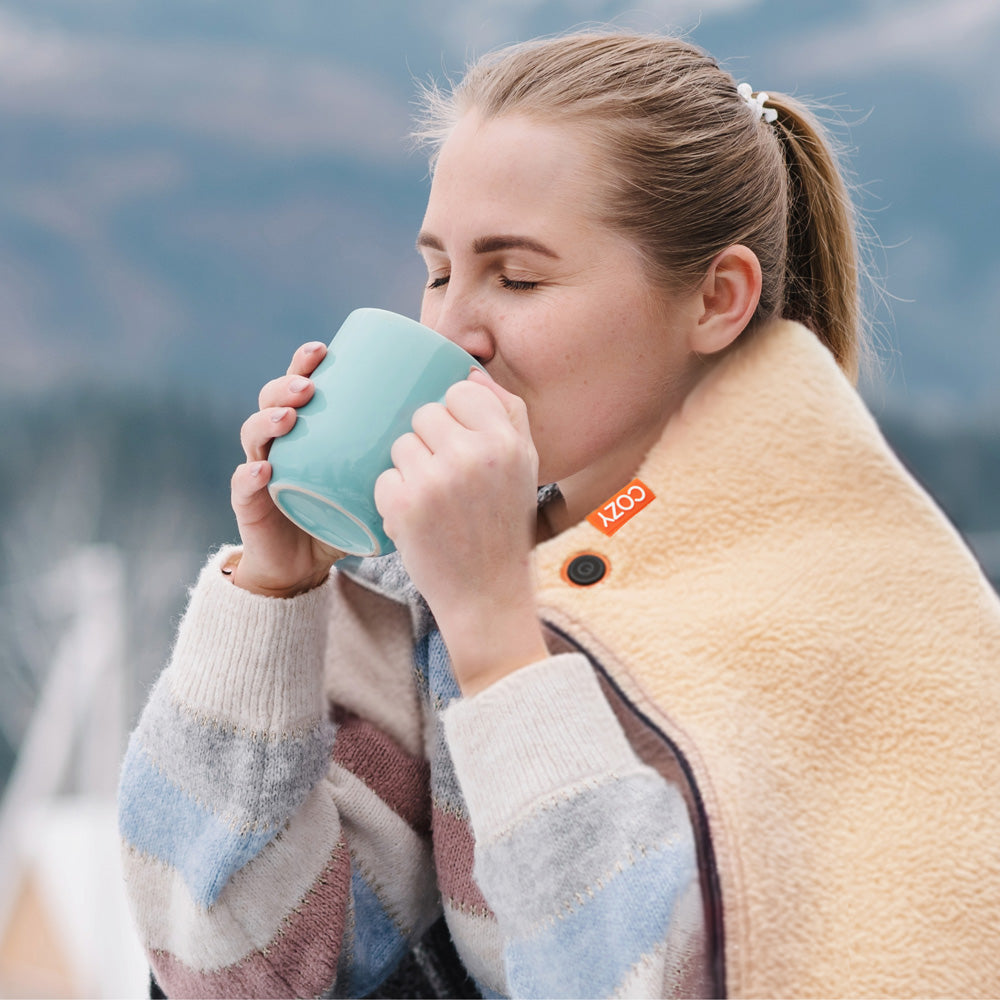A woman, wrapped in the COZY Heated Throw & Seat Cover Pad - JORV and a striped sweater, enjoys a hot drink from a light blue mug outside with blurred mountains in the background.