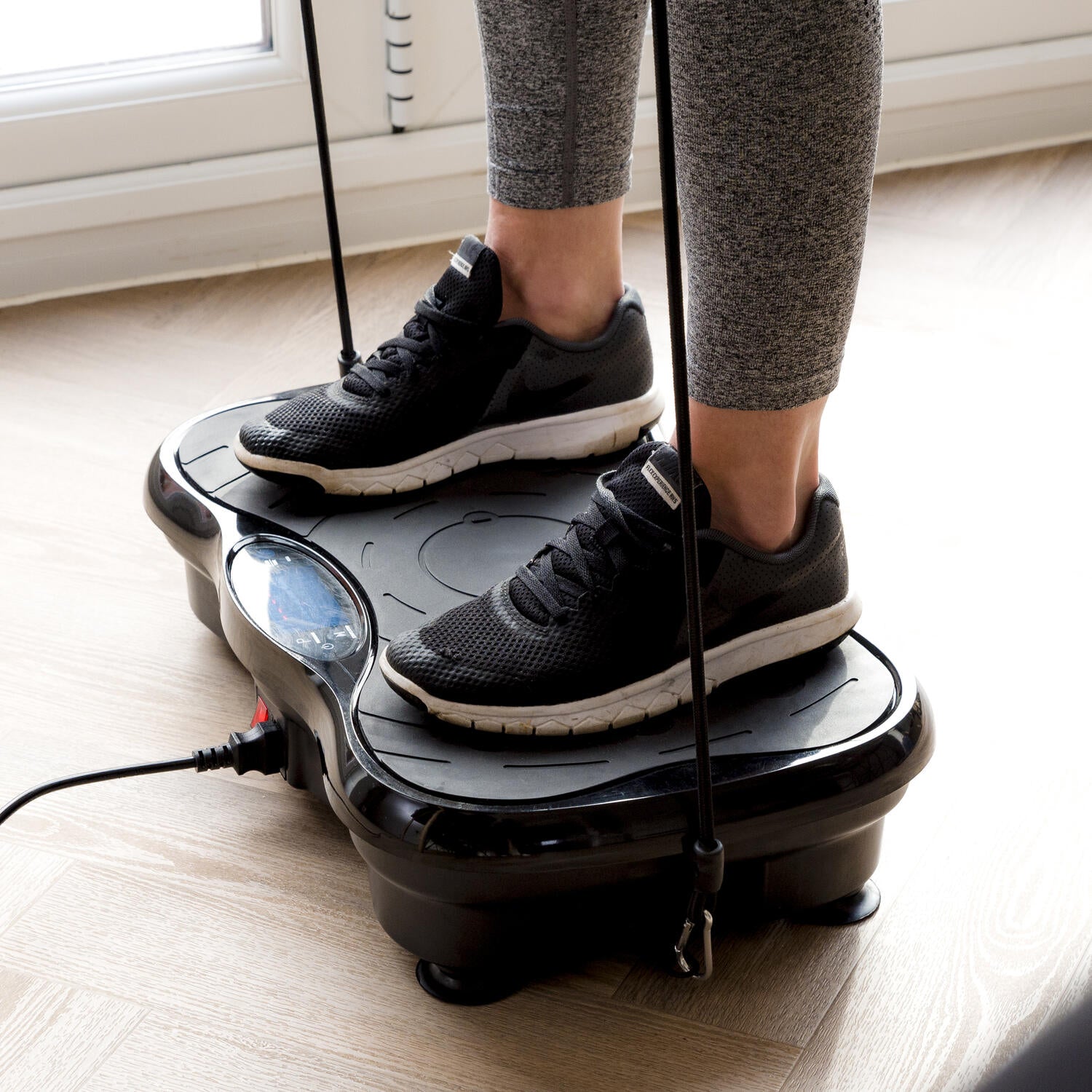 A person stands on the Whole Body Vibration Training Exercise Plate with resistance bands, wearing black sneakers and gray leggings, enjoying a full body indoor workout on a light wood floor.