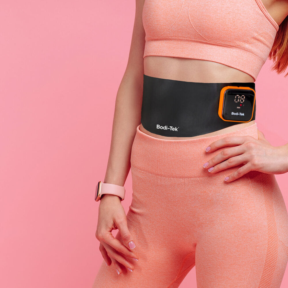 A woman in a peach sports outfit wears a black Ab & Back Muscle Strengthener with heat, featuring a digital display, standing against a pink background.