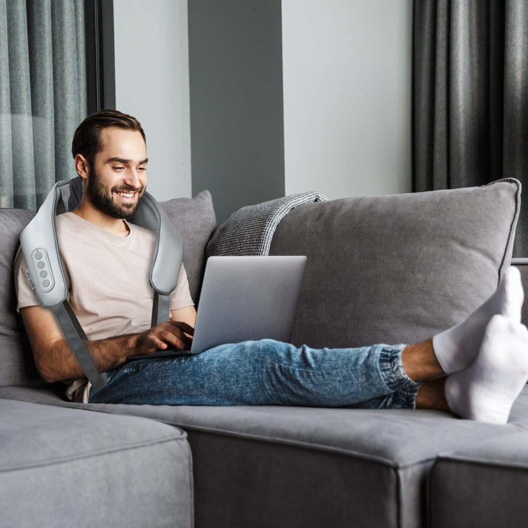 A man sits comfortably on a gray couch using a laptop, smiling and wearing the Ona Neck & Shoulder Massager with infrared heat and control buttons. Gray curtains enhance the modern, relaxed atmosphere of the room.
