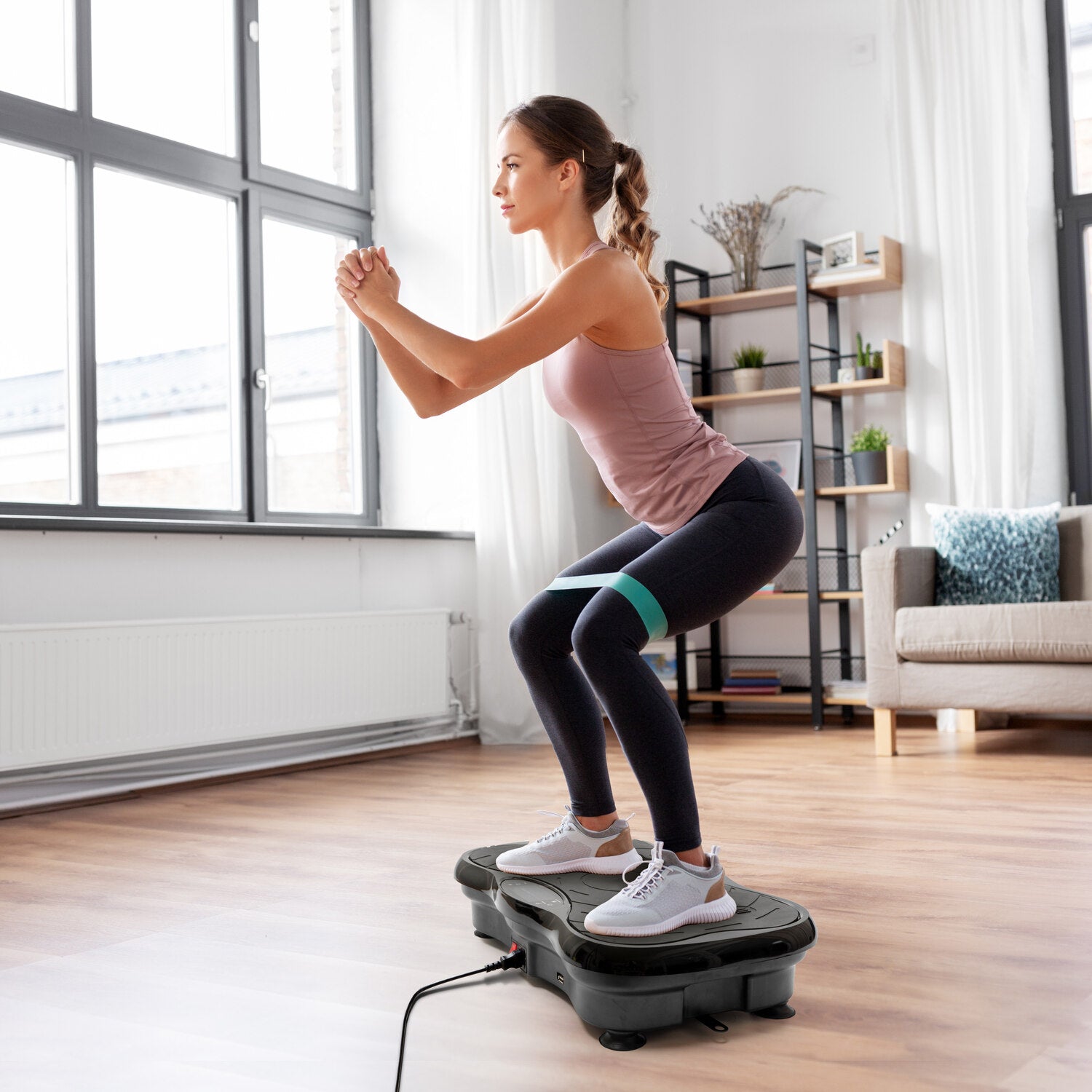 A woman in athletic wear performs squats on the Whole Body Vibration Training Exercise Plate in a bright living room, using a resistance band for a full body workout. Large windows and plant-filled shelves are seen in the background.