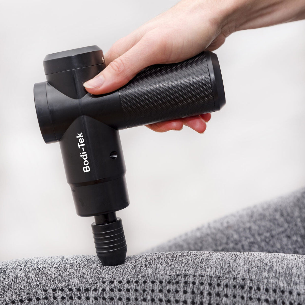 A hand holds a black Hot & Cold Deep Tissue Massager Gun, pressing it against a grey textured fabric surface.