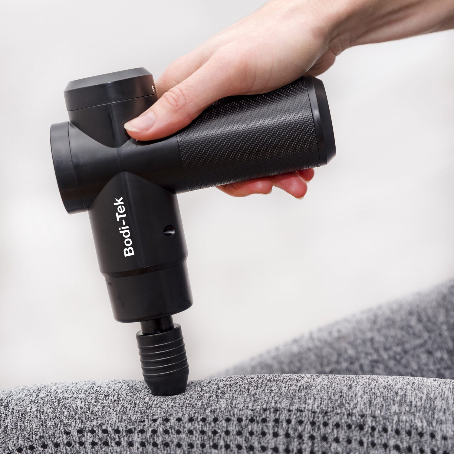 A hand holds a black Hot & Cold Deep Tissue Massager Gun, pressing it against a grey textured fabric surface.