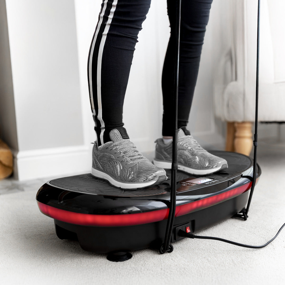 A person in gray sneakers and black leggings with white stripes stands on a 4D Vibration Plate Exercise Machine, ready for a full body workout on the light-carpeted floor.