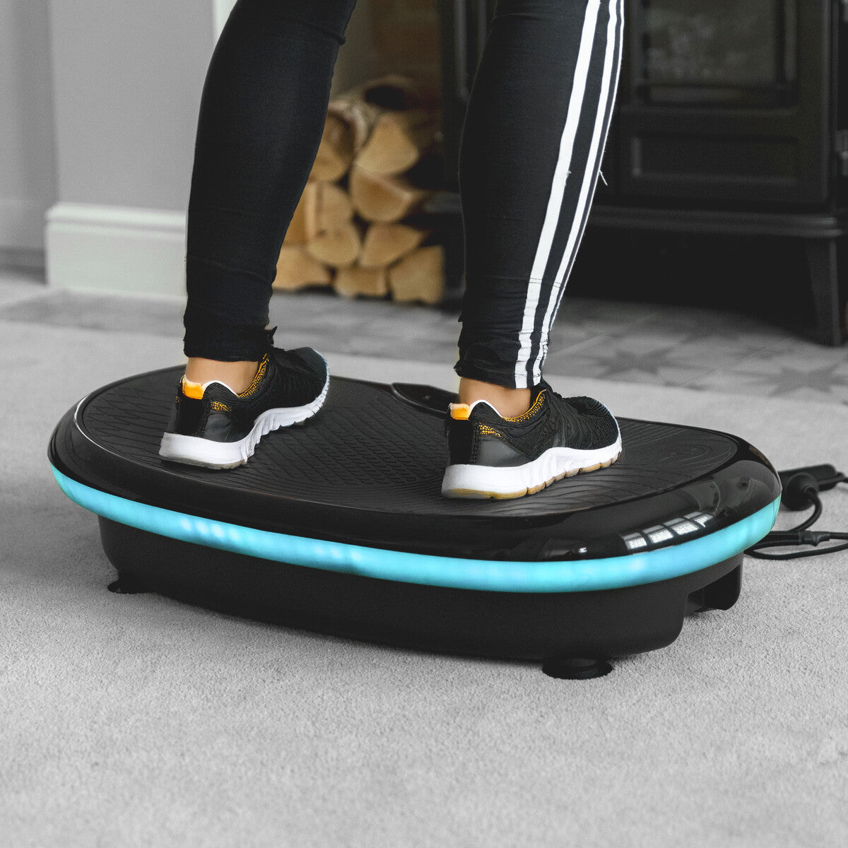 A person in black athletic shoes and striped leggings stands on the 4D Vibration Plate Exercise Machine, designed for full body workouts and fat burning, positioned on a carpeted indoor floor.