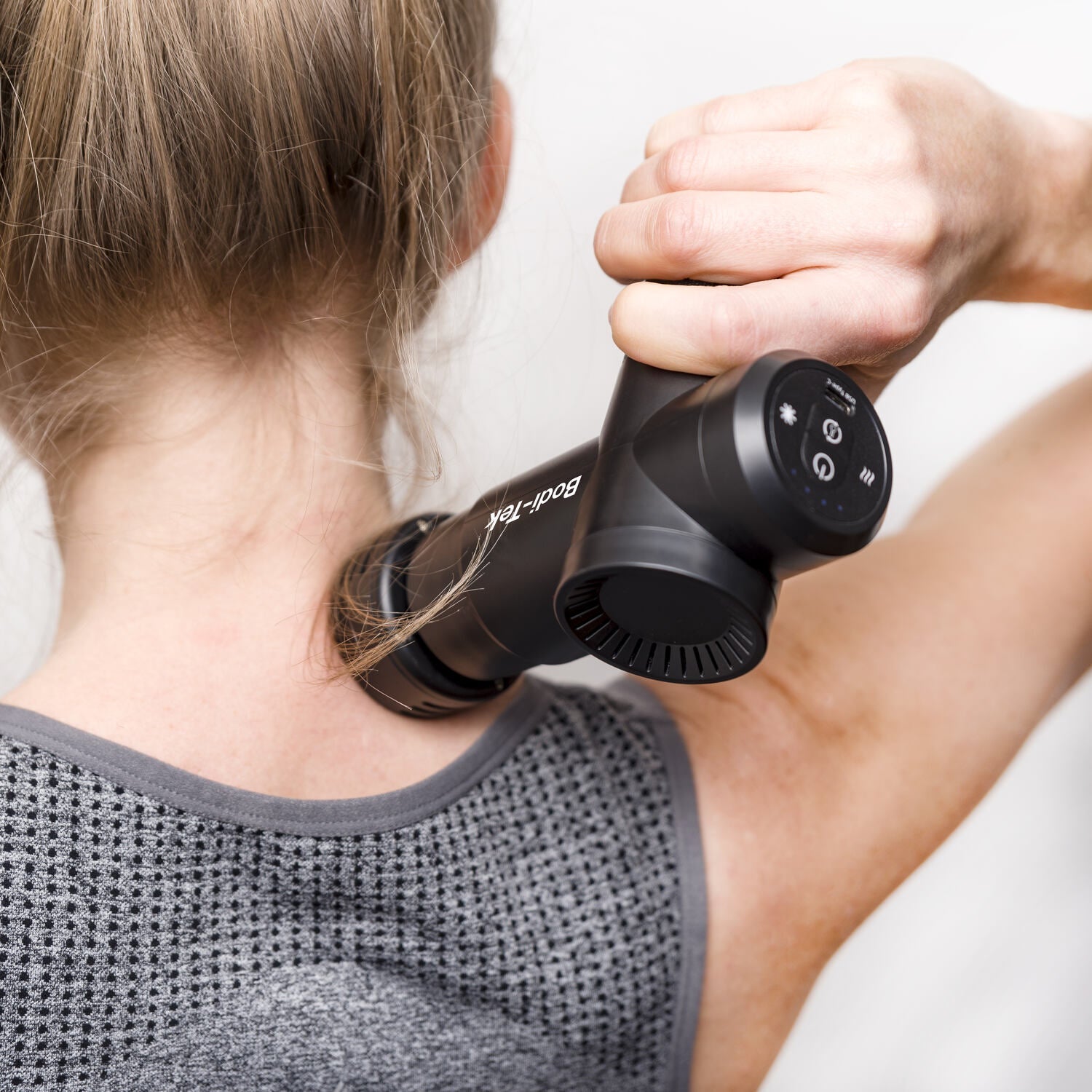 A person uses the Hot & Cold Deep Tissue Massager Gun on their neck and shoulder while wearing a gray athletic top. The devices display and controls are visible, aiding muscle recovery.