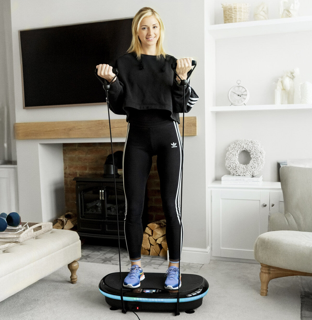 A woman in black athletic wear stands on the 4D Vibration Plate Exercise Machine, using resistance bands for a full body workout in a bright living room with shelves, a fireplace, and stylish home decor.