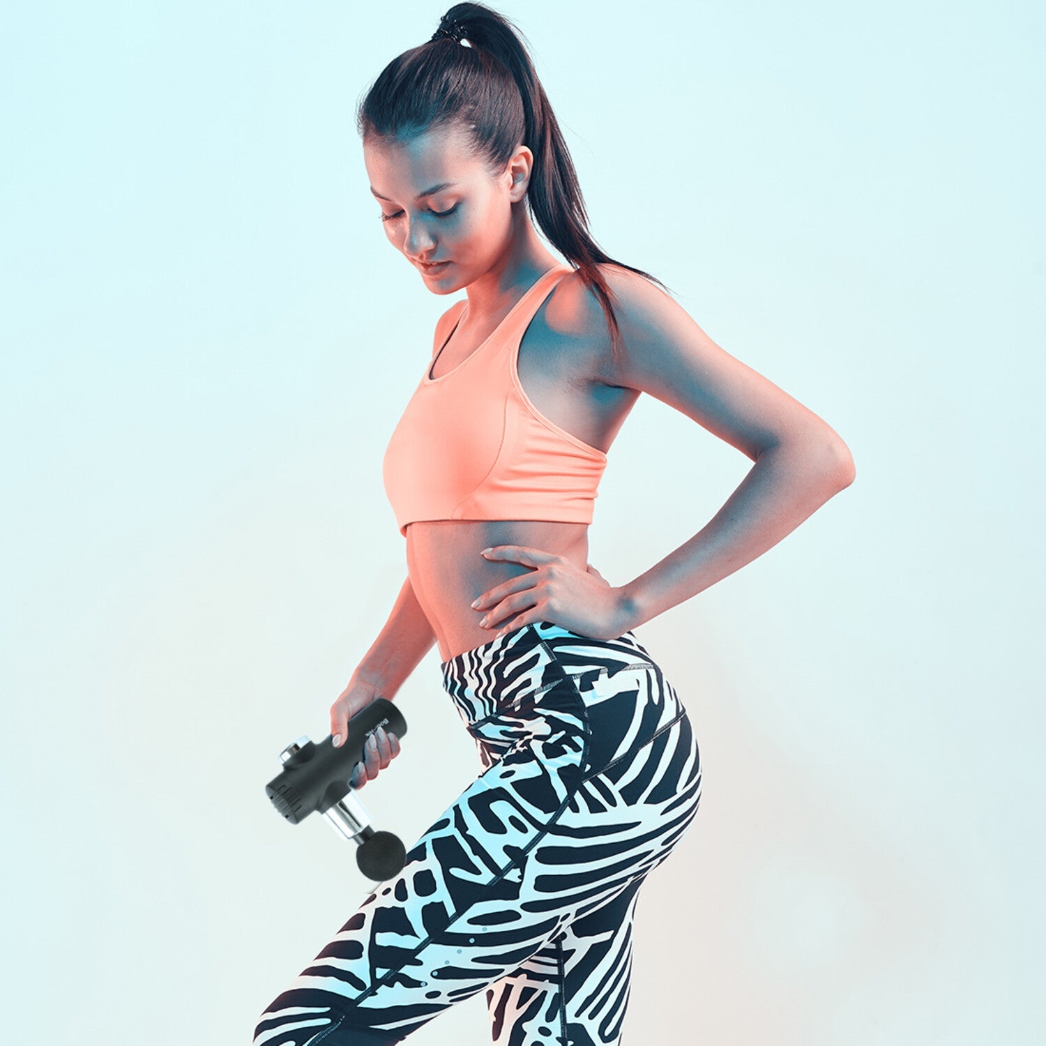 A woman in a peach sports bra and patterned leggings uses the Mini Deep Tissue Massage Gun on her thigh while standing against a light blue background.
