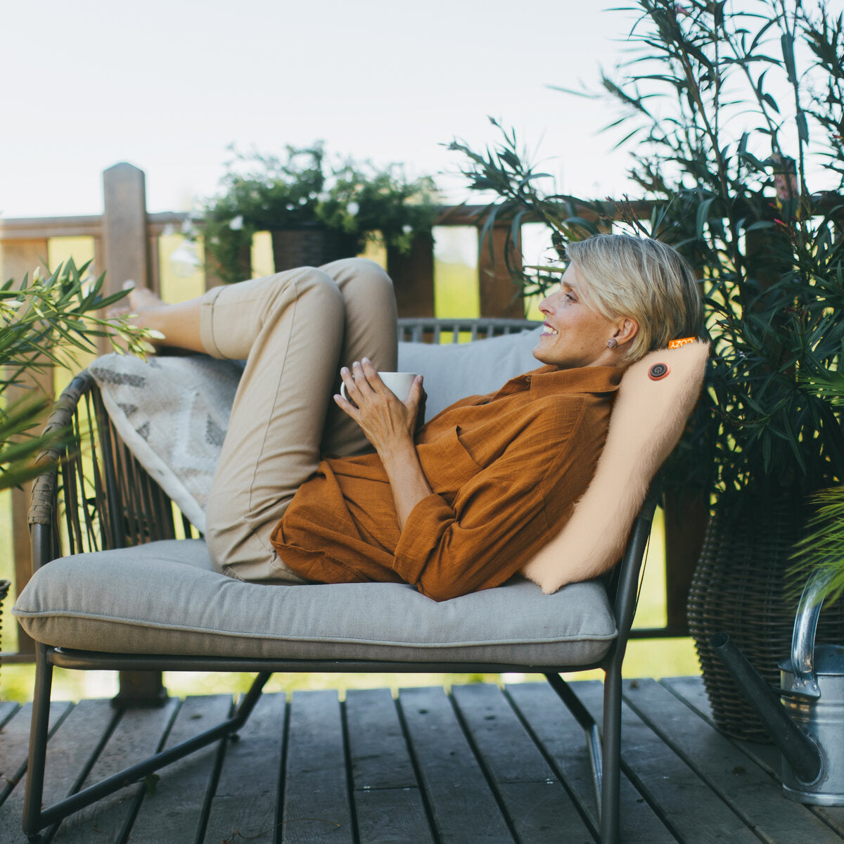 A woman relaxes on an outdoor lounge chair, smiling with a mug in hand among green plants. She rests her head on the COZY Heated Cushion Cordless - UNA Faux Fur (45cm x 45cm) for extra comfort.