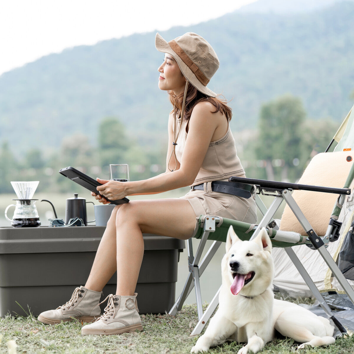 A woman in beige outdoor clothing sits on a COZY Heated Cushion Cordless - UNA Knitted (45cm x 45cm) on a camping chair, holding a tablet. A coffee setup, glass, and white dog are nearby, with a tent and green hills in the background.