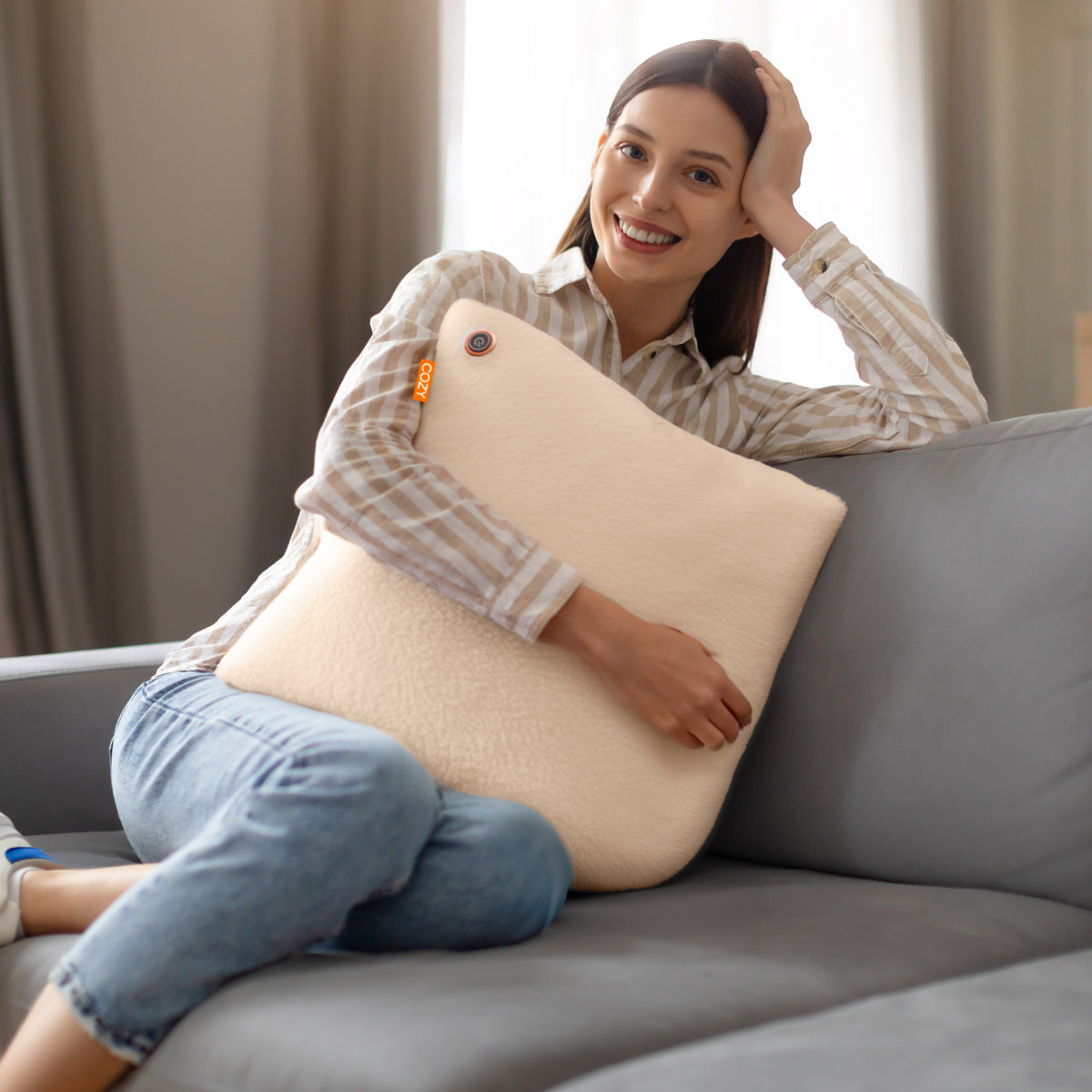 A woman in a striped shirt and jeans sits on a gray sofa, smiling as she hugs the COZY Heated Cushion Cordless - UNA Faux Fur (45cm x 45cm) in beige, enjoying its soft warmth with one arm resting on the sofa’s back.