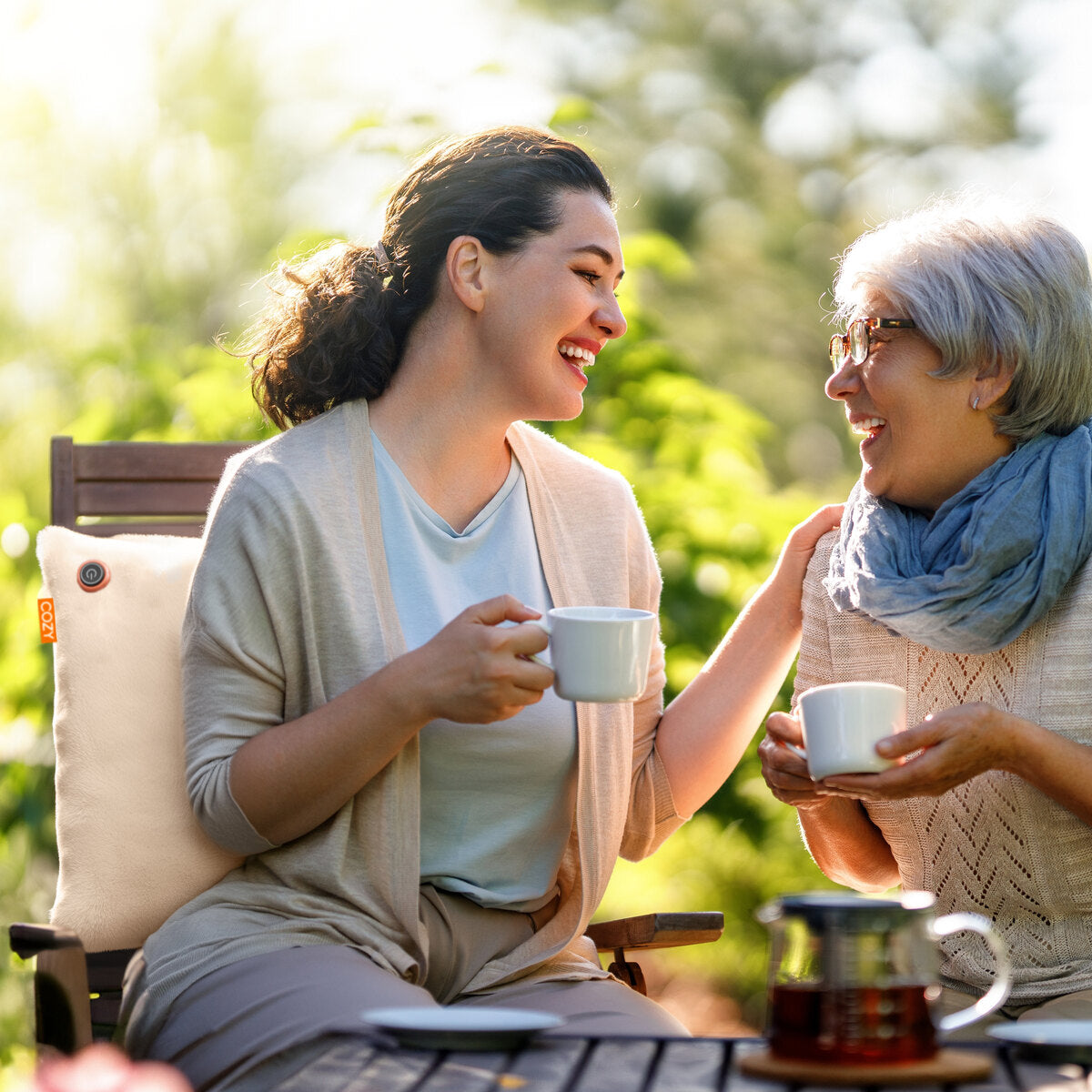 Two women sit outdoors on wooden chairs, smiling with white mugs in hand. The COZY Heated Cushion Cordless - UNA Faux Fur (45cm x 45cm) adds warmth and comfort to their cheerful, sunlit gathering surrounded by greenery.