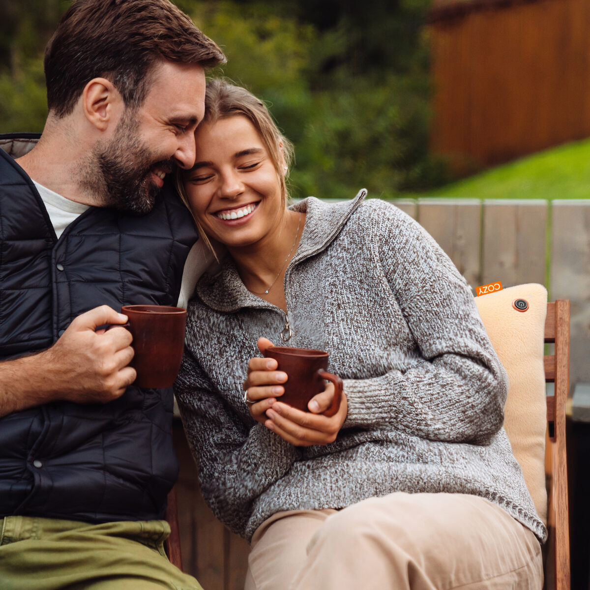 A smiling couple sits outdoors, happily leaning together with mugs in hand, staying warm and relaxed on the COZY Heated Cushion Cordless - UNA Woolly (45cm x 45cm), dressed in cozy sweaters.