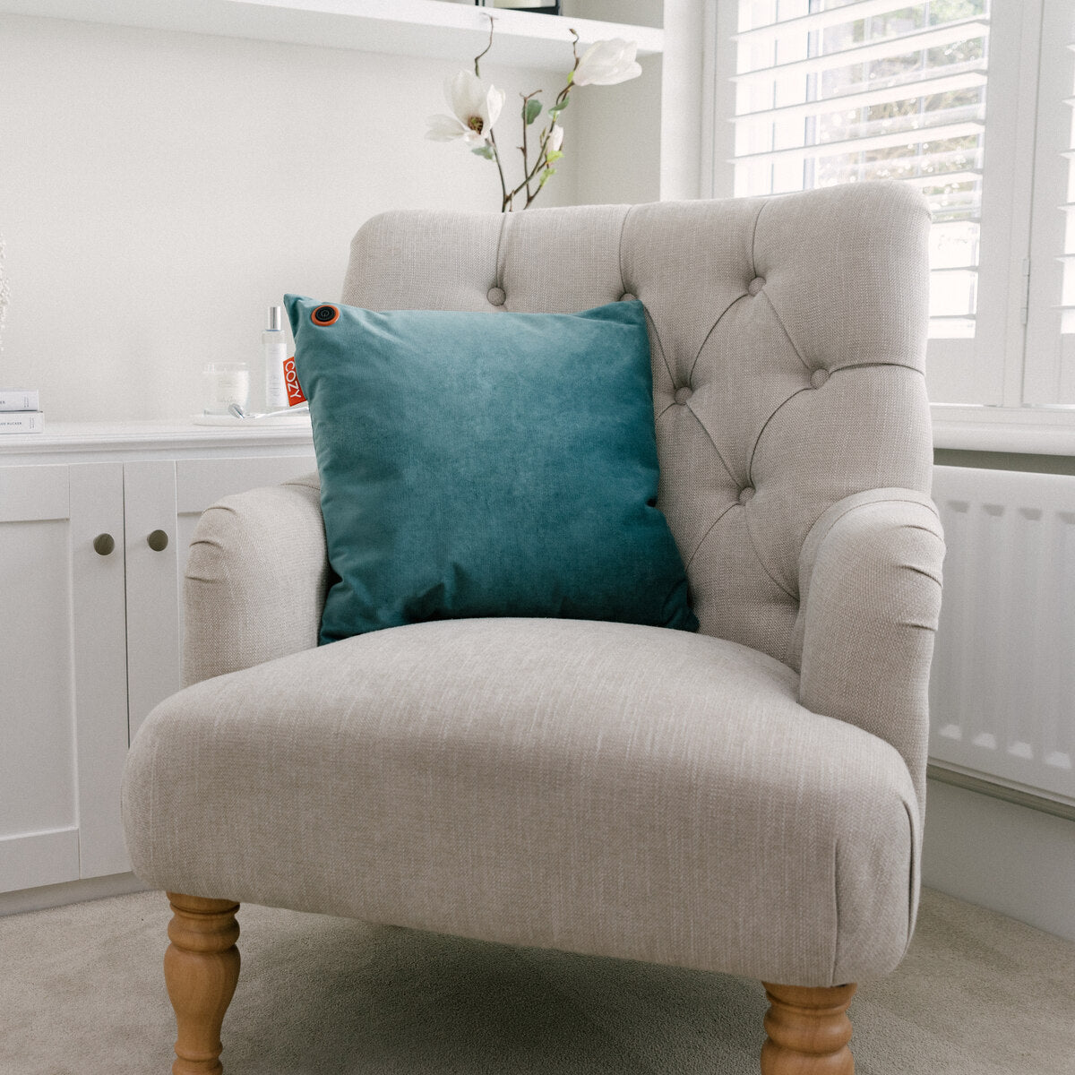 A light beige upholstered armchair with wooden legs features the COZY Heated Cushion Cordless - UNA Velvet (45cm x 45cm) in teal. The chair sits in a bright room with white cabinets, a window with shutters, and a white orchid.