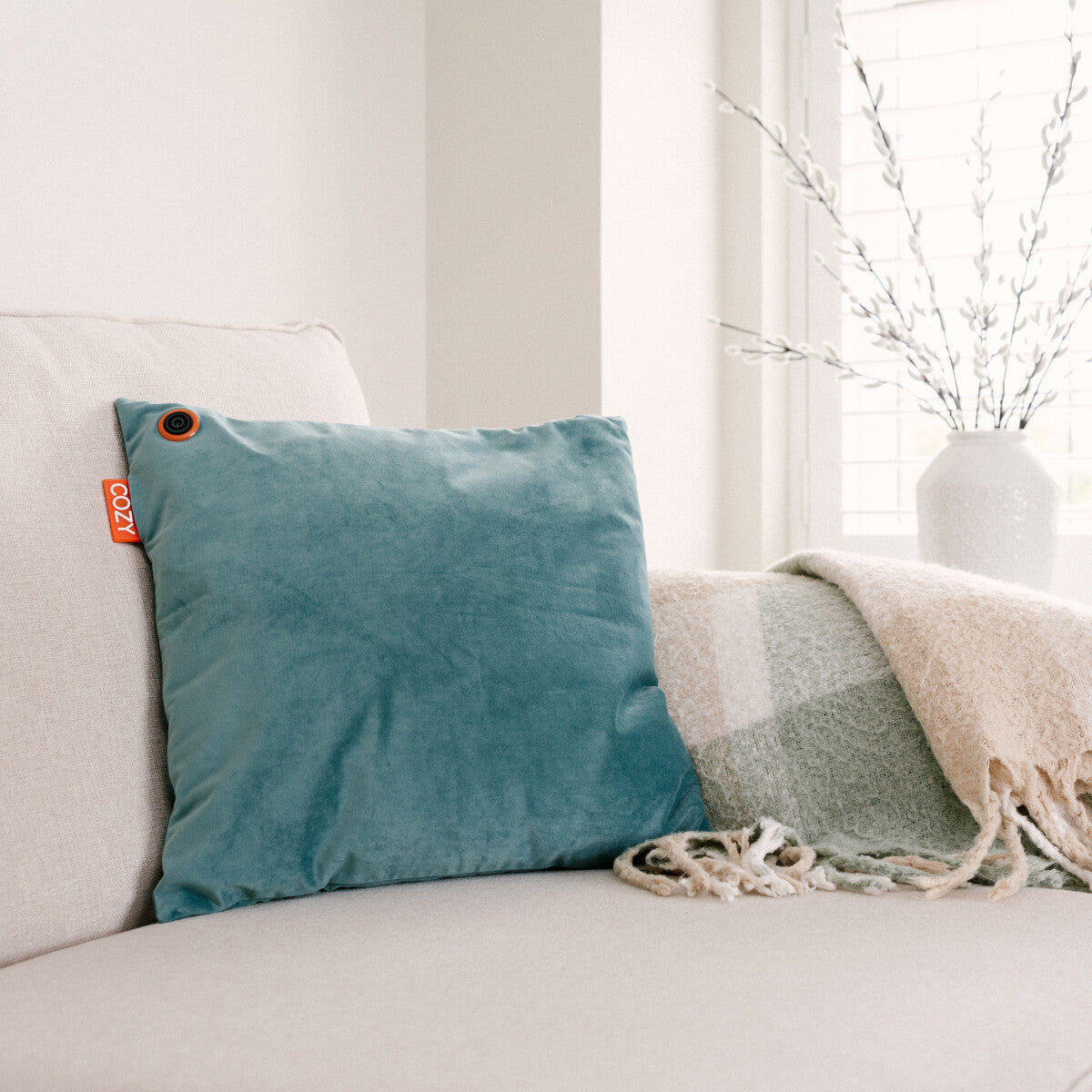 A COZY Heated Cushion Cordless - UNA Velvet (45cm x 45cm), featuring a teal cover with an orange label and button, rests on a light beige sofa beside a green and cream plaid throw blanket. A white vase with branches sits on the windowsill behind.