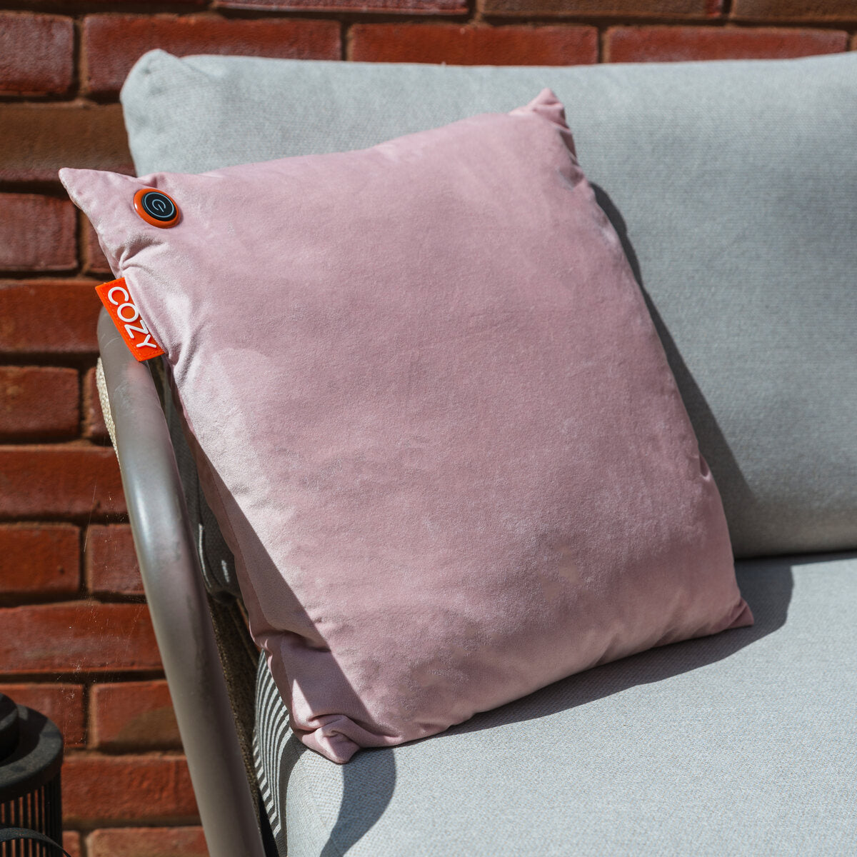 A COZY Heated Cushion Cordless - UNA Velvet (45cm x 45cm) in light pink with a COZI tag and top-corner button sits on a light gray outdoor sofa against a red brick wall.