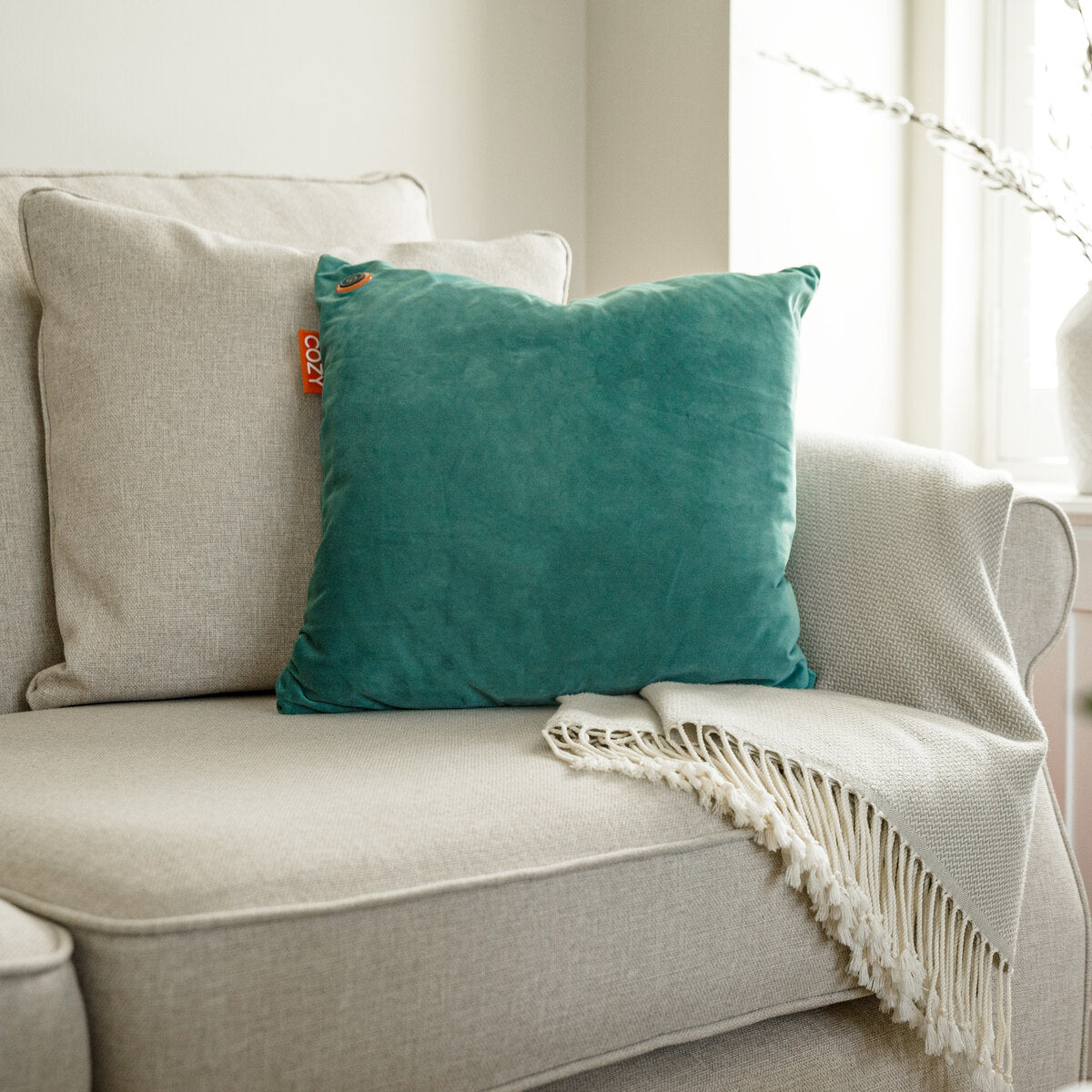 A COZY Heated Cushion Cordless - UNA Velvet (45cm x 45cm) in teal sits on a light beige sofa with a textured cream blanket by a sunlit window.