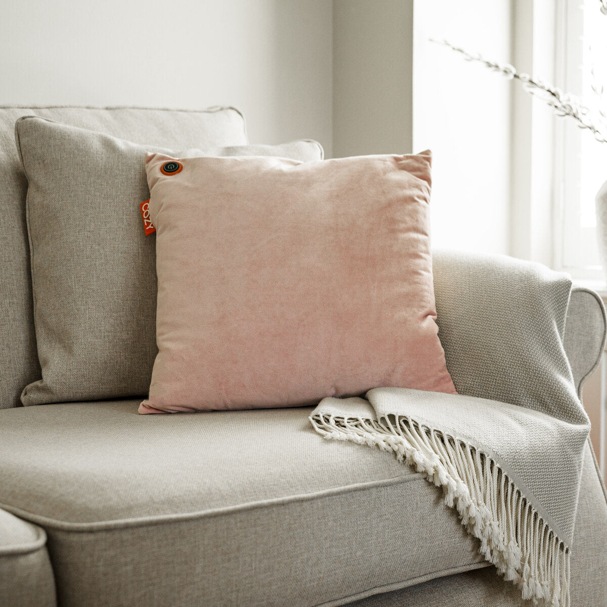 A COZY Heated Cushion Cordless - UNA Velvet (45cm x 45cm) in light pink sits on a beige textured sofa. A fringed light gray blanket covers the arm, and soft natural light flows in from a nearby window.