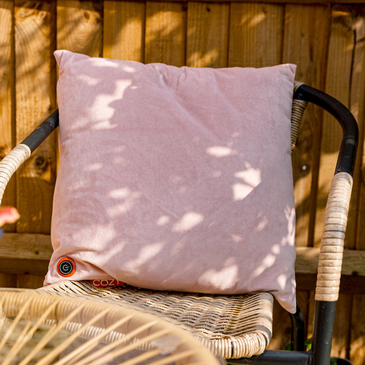 The COZY Heated Cushion Cordless - UNA Velvet (45cm x 45cm) in pale pink rests on a rattan chair outdoors, sunlight filtering through a wooden fence, its small circular black and red logo visible.