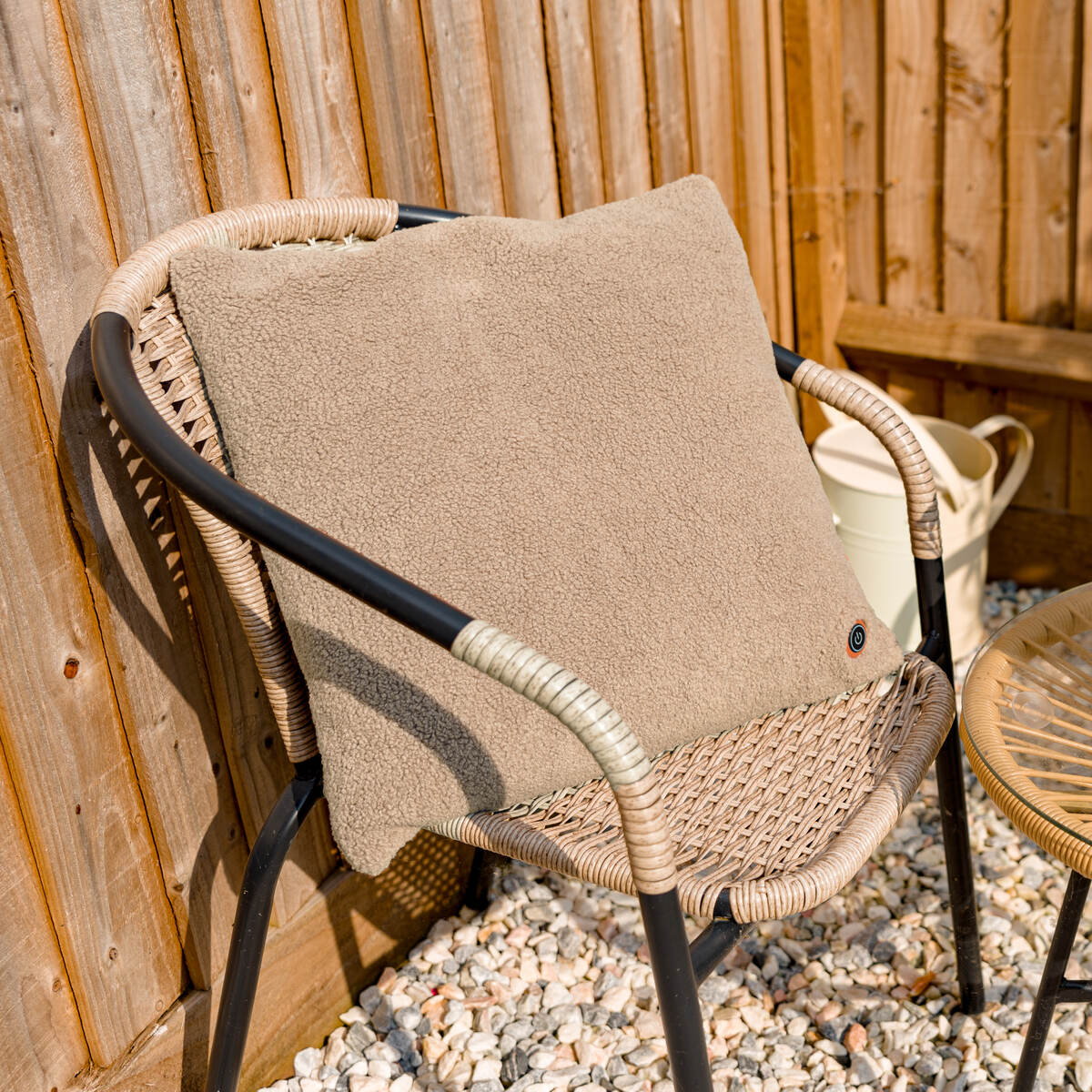 A COZY Heated Cushion Cordless - UNA Woolly (45cm x 45cm), in beige, sits on a wicker chair with black metal arms on pebble ground by a wooden fence. A white watering can and a small table are placed nearby.