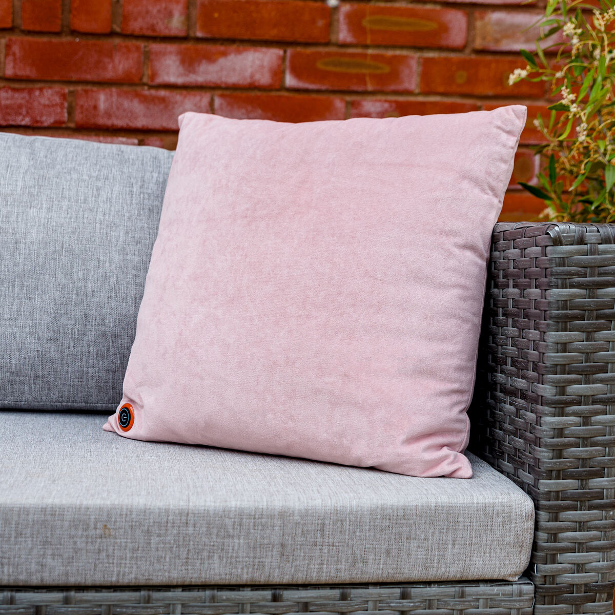 A light pink COZY Heated Cushion Cordless - UNA Velvet (45cm x 45cm) sits on a gray outdoor wicker sofa, with a red brick wall and green plant behind. A small circular patch accents the bottom corner of the cushion.