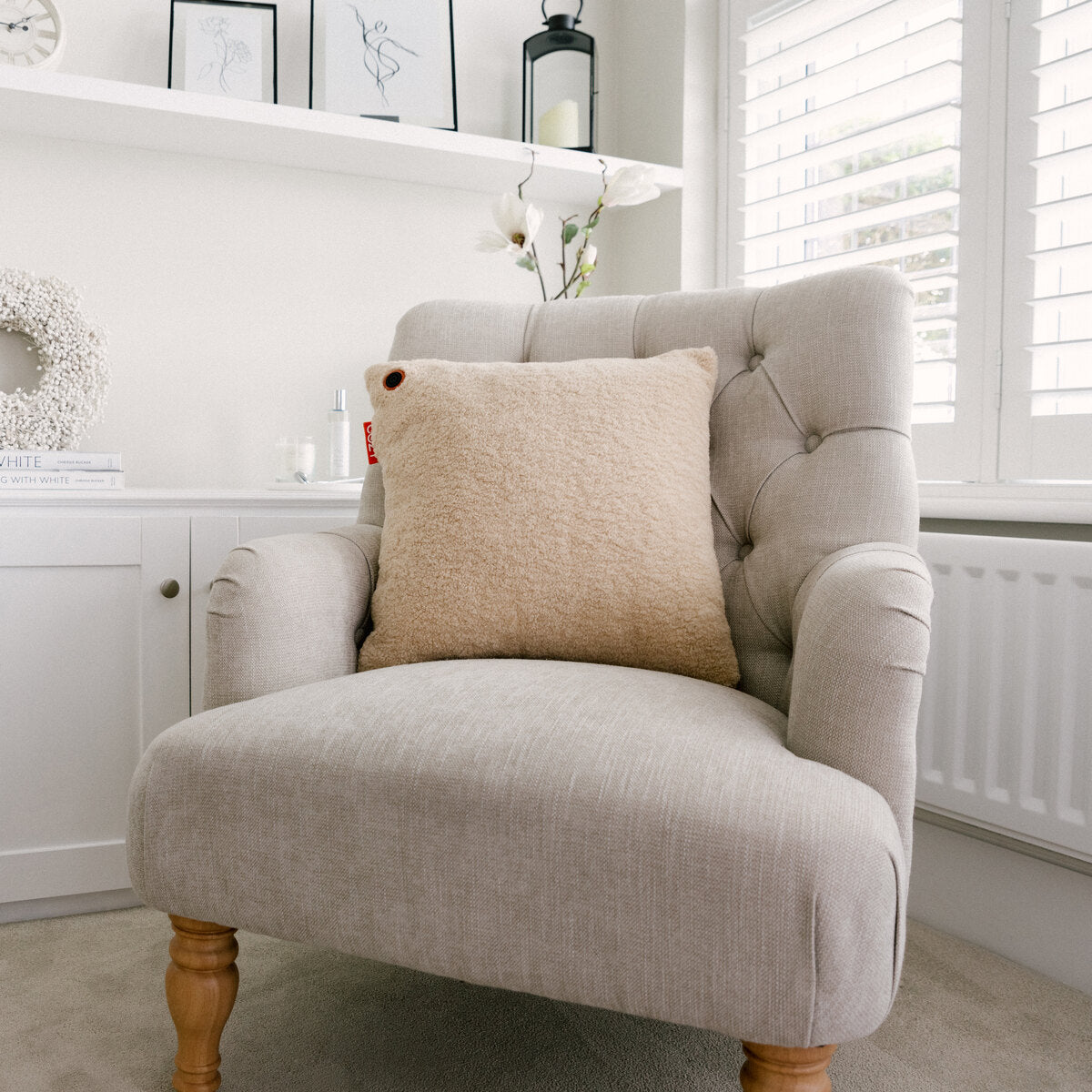 A light grey upholstered armchair with wooden legs and a COZY Heated Cushion Cordless - UNA Woolly (45cm x 45cm) rests in a bright, cozy room with white walls, a shuttered window, and minimal decor on a shelf behind it.