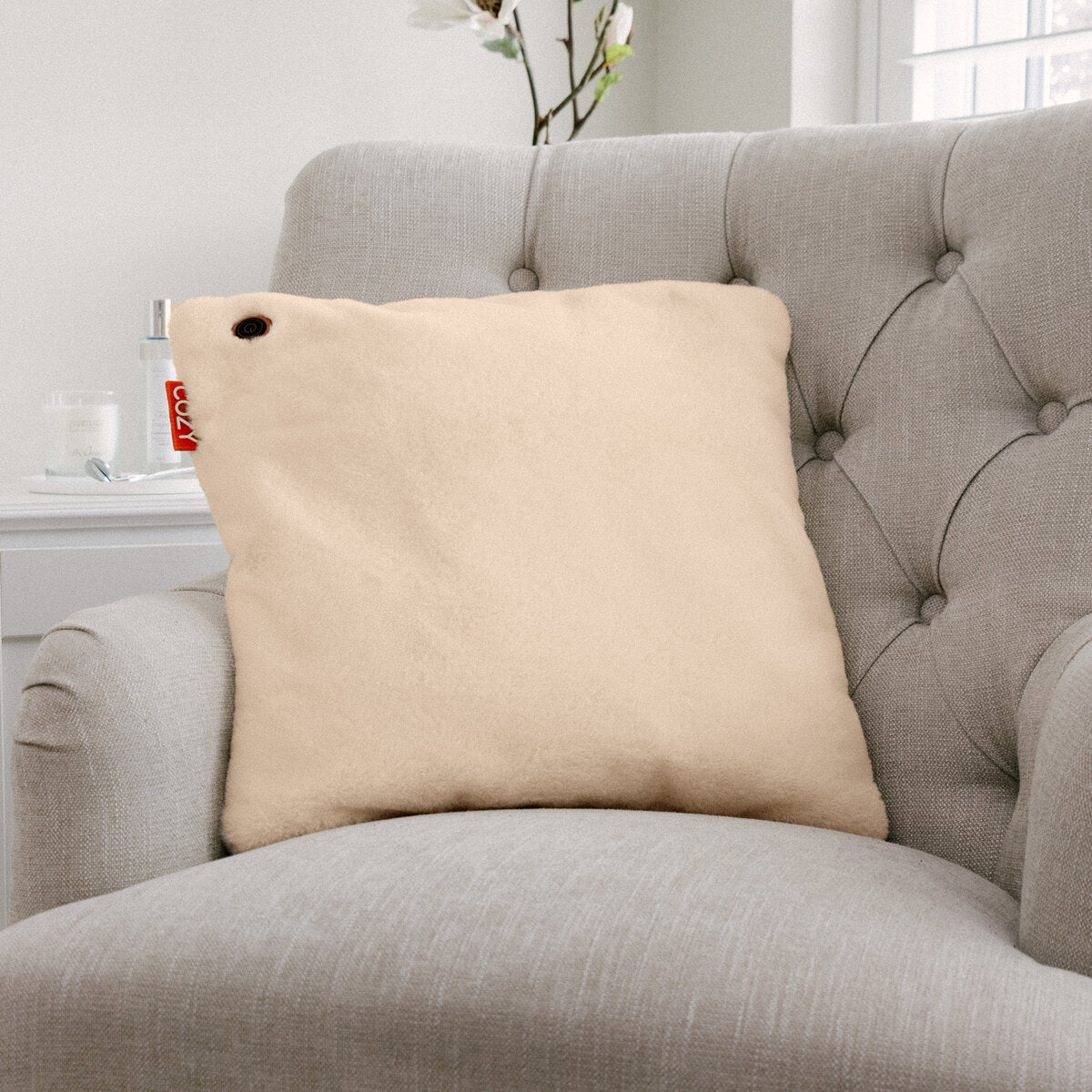 The COZY Heated Cushion Cordless - UNA Faux Fur (45cm x 45cm), a light beige square cushion with a small red tag and dark button, sits on a light gray tufted armchair in a bright room with a vase and window behind.