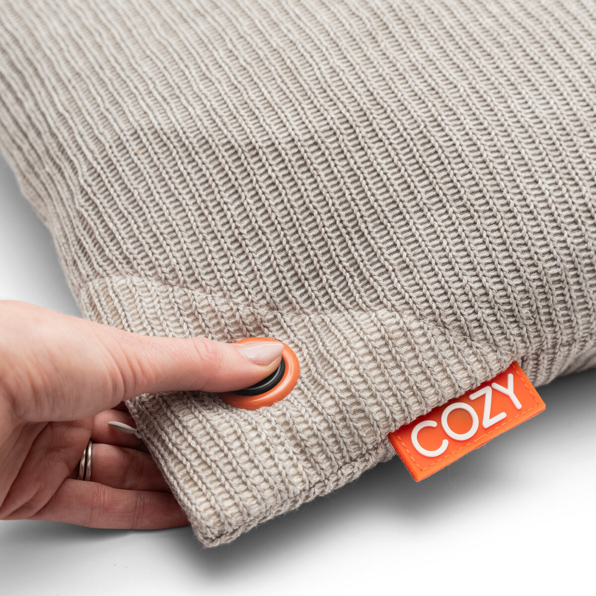 A hand presses a button on the COZY Heated Cushion Cordless - UNA Knitted (45cm x 45cm), featuring a textured beige design and bright orange COZY tag, highlighting comfort and modern style.