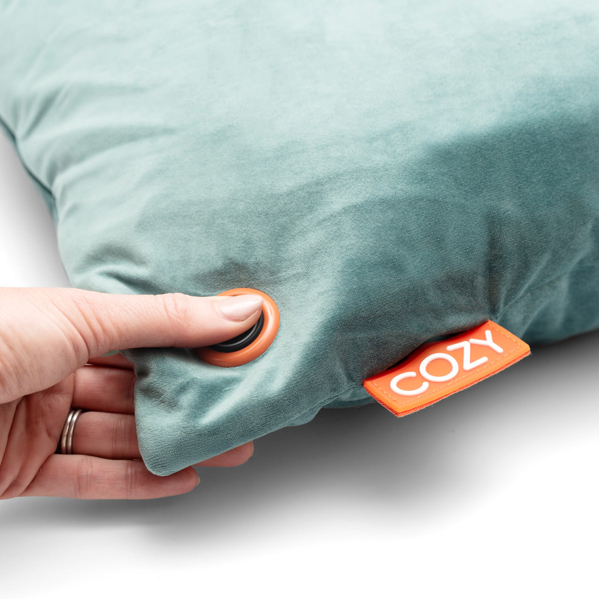 A hand presses a round button on the COZY Heated Cushion Cordless - UNA Velvet (45cm x 45cm), a soft, light teal wireless heated cushion with an orange COZY tag.
