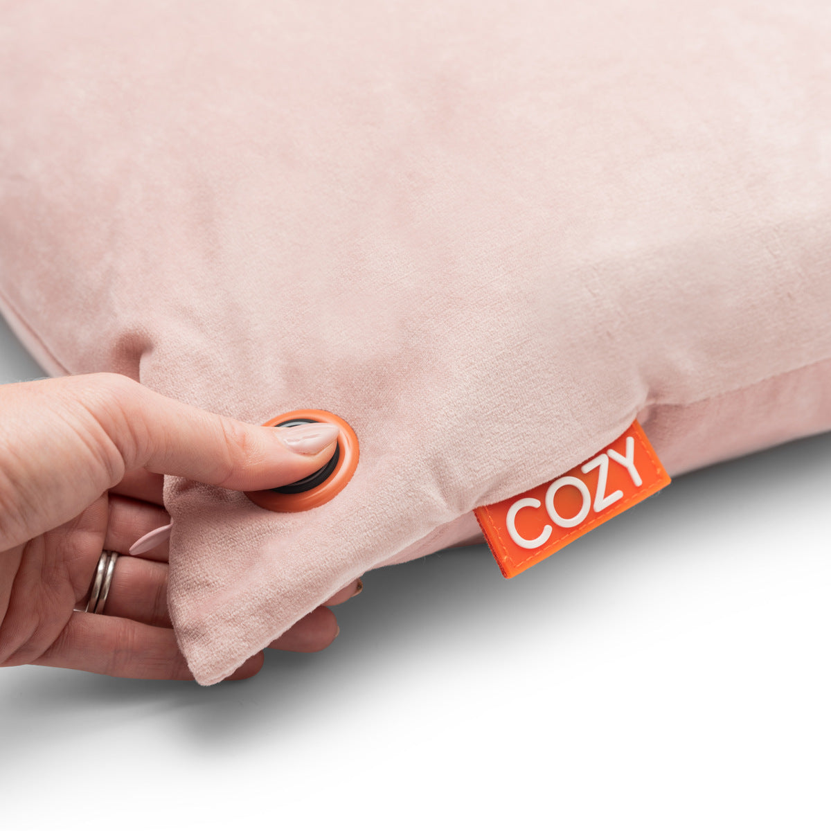 A hand presses a round button on the corner of a soft, light pink COZY Heated Cushion Cordless - UNA Velvet (45cm x 45cm) with an orange tag labeled COZY.