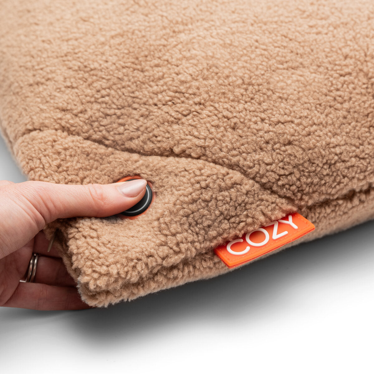 A hand presses a black button on the COZY Heated Cushion Cordless - UNA Woolly (45cm x 45cm), a soft tan fleece cushion with an orange COZY tag—perfect for warmth and comfort anywhere.