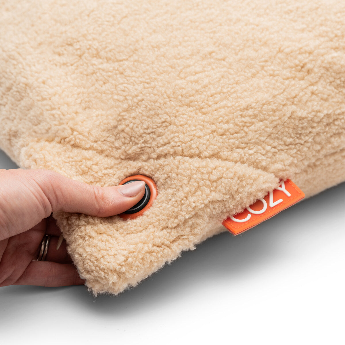 A hand presses a black button on the soft beige UNA Woolly (45cm x 45cm) COZY Heated Cushion Cordless, with a visible orange COZY tag—revealing it’s actually a portable heated cushion.