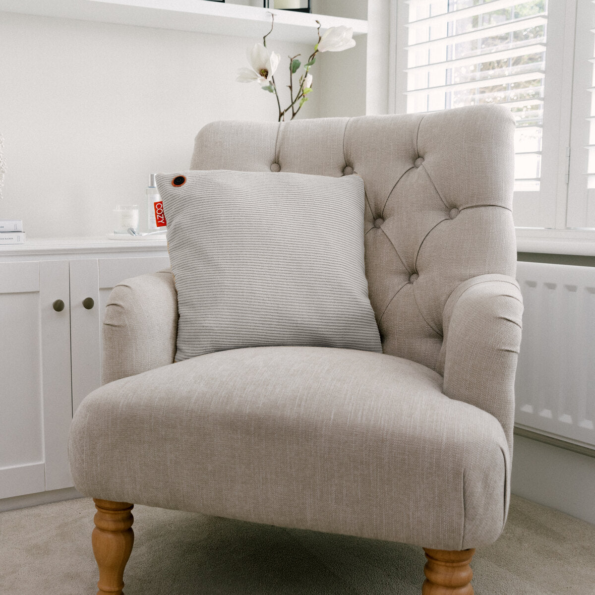 A light beige upholstered armchair with wooden legs and a COZY Heated Cushion Cordless - UNA Knitted (45cm x 45cm) sits in a bright room near white cabinets, a radiator, and a window with white shutters. A white orchid is visible in the background.