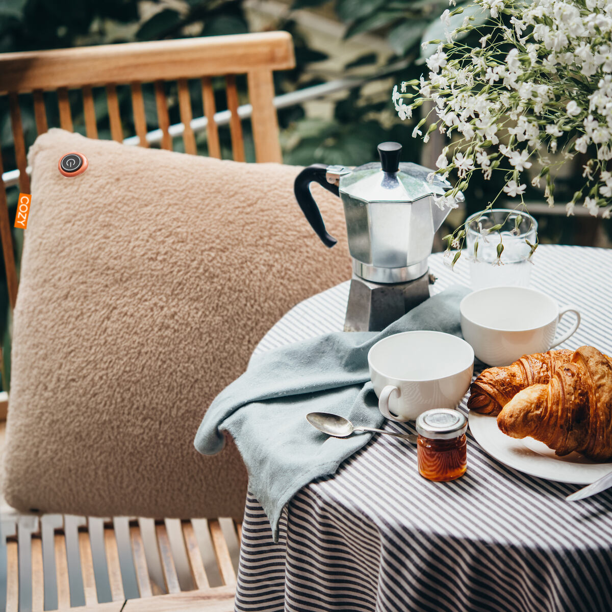 A cozy outdoor breakfast with a striped tablecloth, coffee pot, white cups, croissants, jam jar, white flowers, and the COZY Heated Cushion Cordless - UNA Woolly (45cm x 45cm) warming a wooden chair.