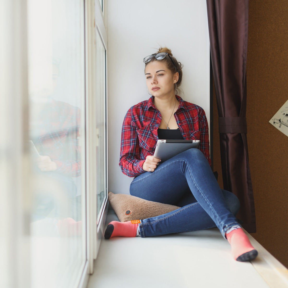 A woman in a red plaid shirt and jeans sits on a windowsill, holding a tablet and relaxing against the COZY Heated Cushion Cordless - UNA Woolly (45cm x 45cm) for extra comfort while looking thoughtfully outside.