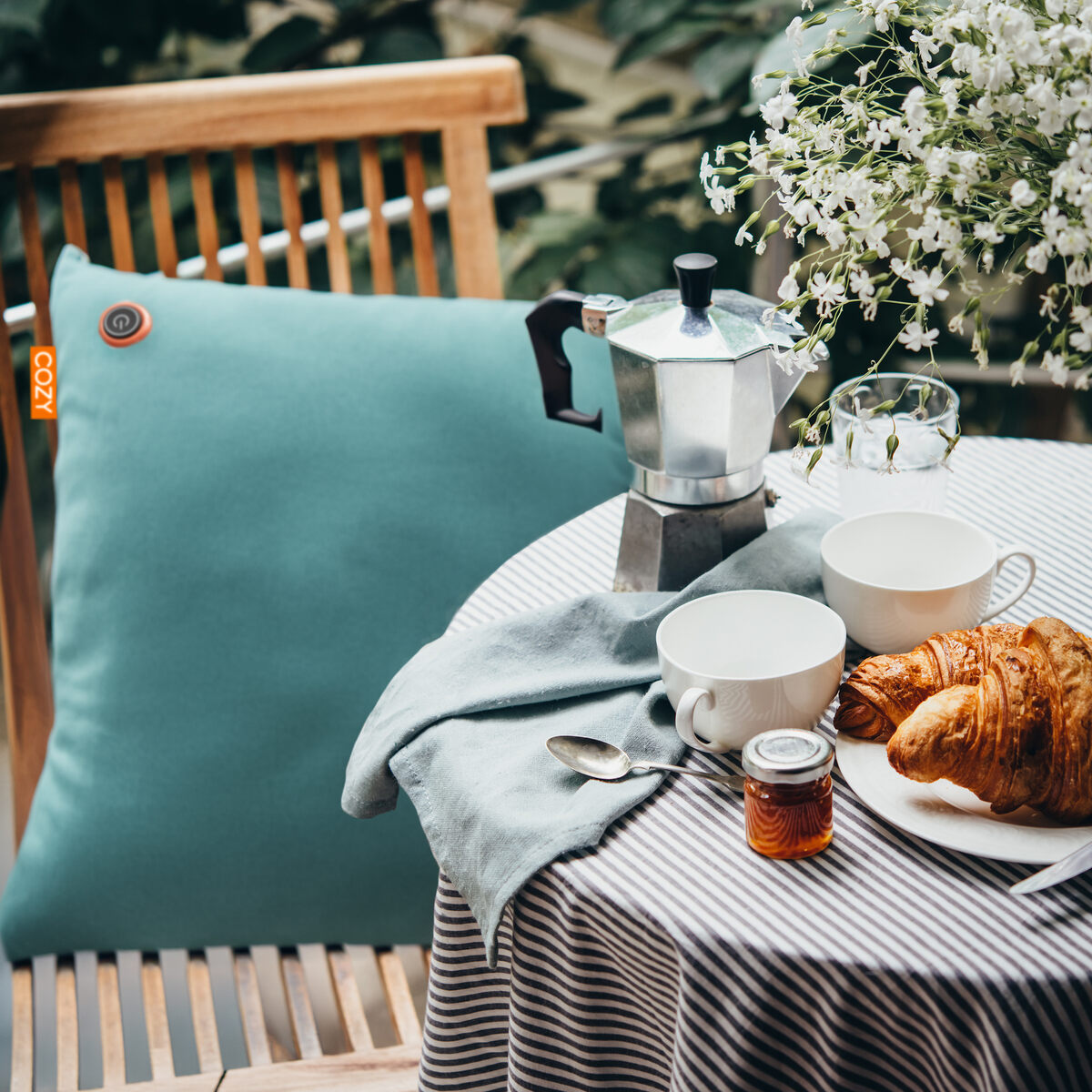 A striped table displays a croissant, jam jar, two white cups, moka pot, and glass of water; the COZY Heated Cushion Cordless - UNA Velvet (45cm x 45cm) sits on a wooden chair beside the table, with white flowers in the background.