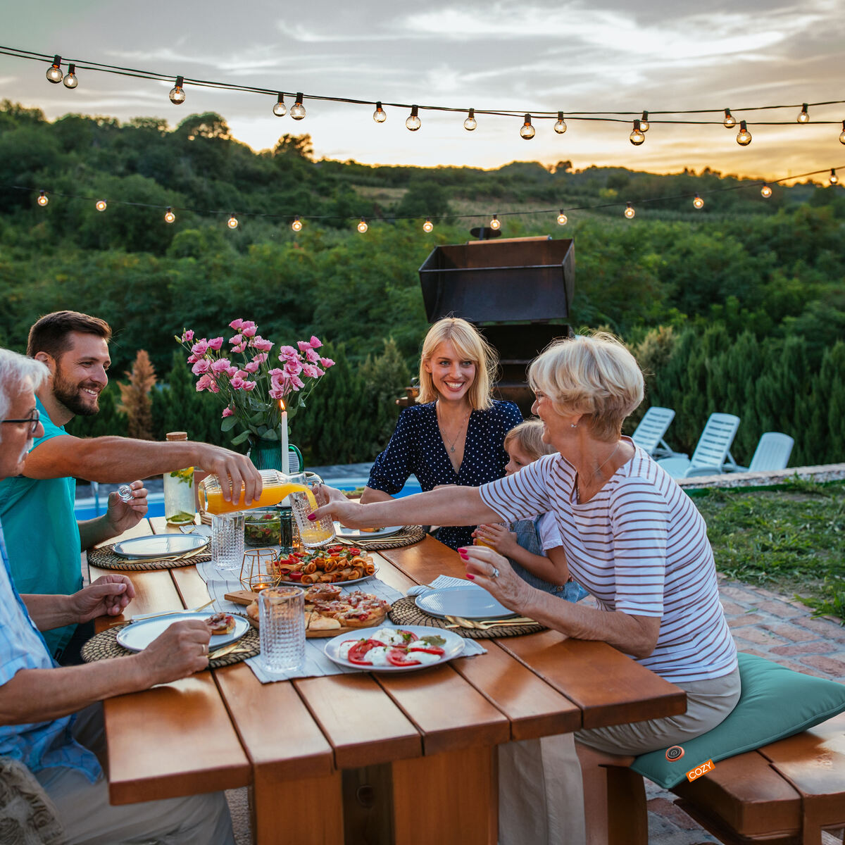 A group of adults and a child share a meal at sunset on a flower-decorated wooden outdoor table, surrounded by greenery and string lights, relaxing on COZY Heated Cushion Cordless - UNA Velvet (45cm x 45cm) wireless cushions.