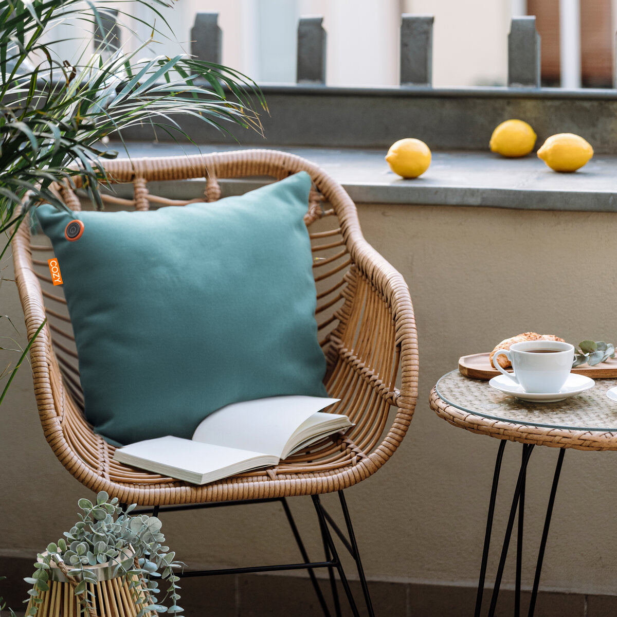 A wicker chair topped with a COZY Heated Cushion Cordless - UNA Velvet (45cm x 45cm) and an open book sits beside a round table holding coffee, a saucer, and a plant. Three lemons are on the background ledge.