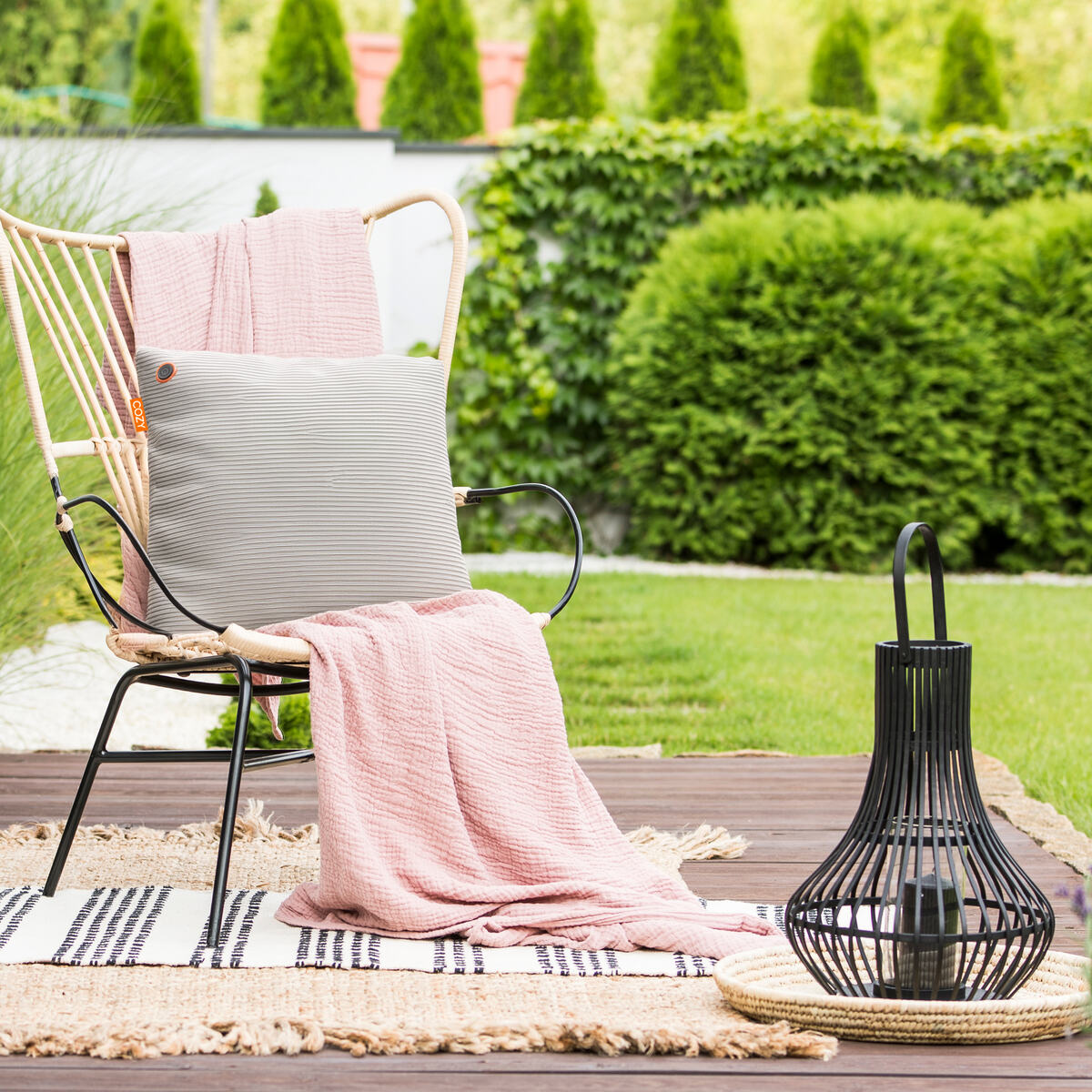 A cozy patio features a chair with a pink blanket, gray pillow, and the COZY Heated Cushion Cordless - UNA Knitted (45cm x 45cm), set on a patterned rug next to a black lantern. Green bushes and trees complete the tranquil outdoor scene.