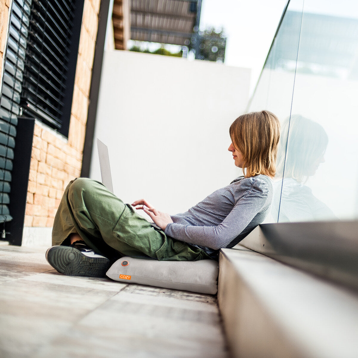 A woman uses her laptop while sitting on the COZY Heated Cushion Cordless - UNA Knitted (45cm x 45cm) in a modern, bright space. She wears a striped long-sleeve shirt and green pants, enjoying warmth and comfort in her cozy work spot.