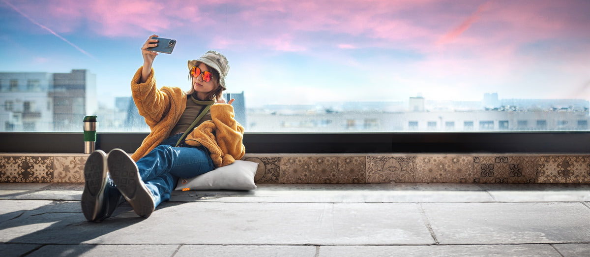 A person in a yellow jacket, jeans, and bucket hat sits on the COZY Heated Cushion Cordless - UNA Knitted (45cm x 45cm) by a large window, taking a selfie with a travel mug nearby and a sunset cityscape in the background.