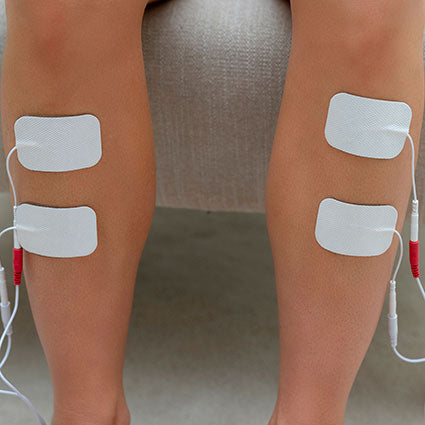 A close-up of two legs with Reviber Premium Electrode Gel Pads (x2) attached to the calves, connected by wires for electrical muscle stimulation therapy. The person is seated on a light-colored surface.