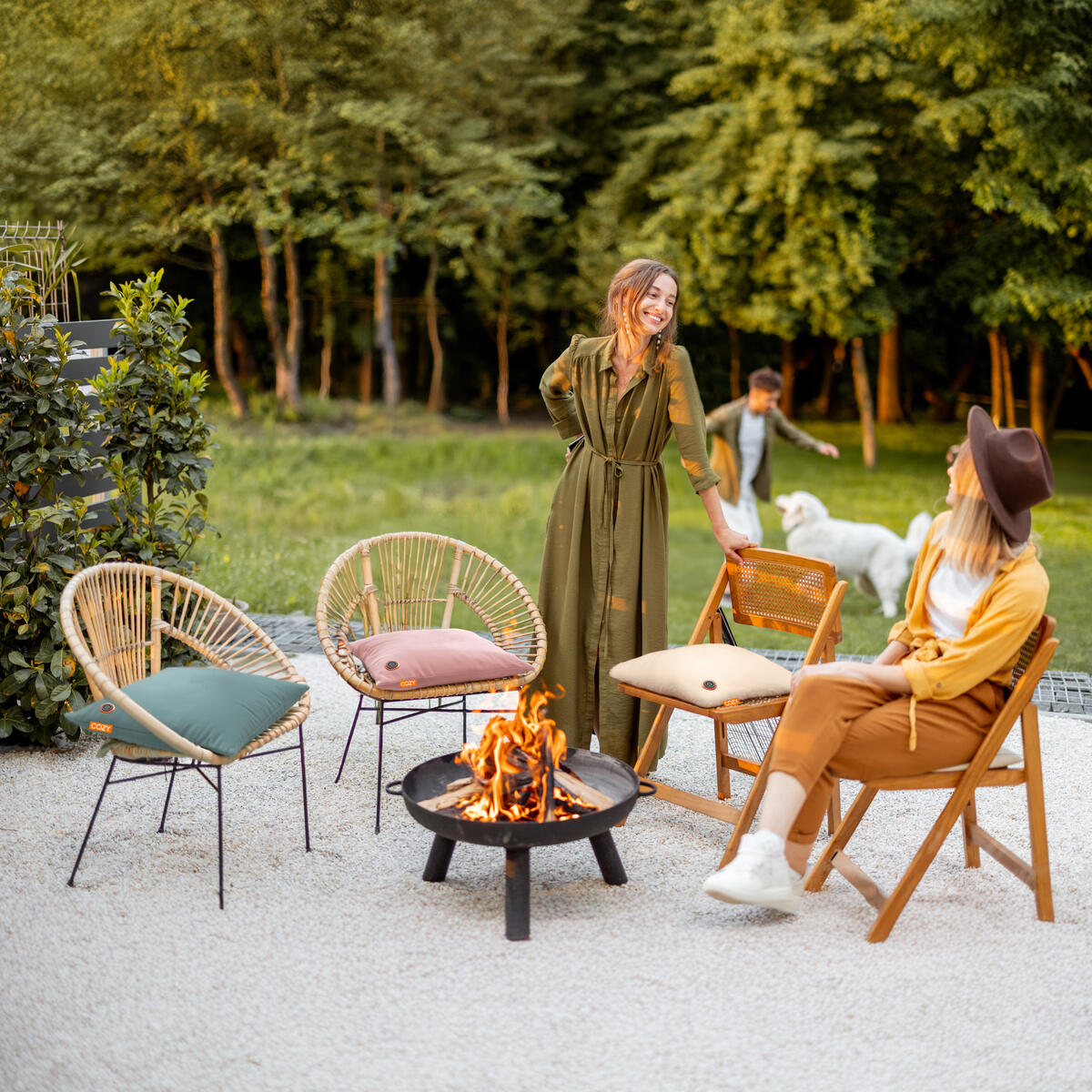 Two women chat beside a fire pit on the patio, relaxing in chairs featuring COZY Heated Cushion Cordless - UNA Velvet (45cm x 45cm), as a child and white dog play nearby. Lush trees and greenery surround the welcoming outdoor space.