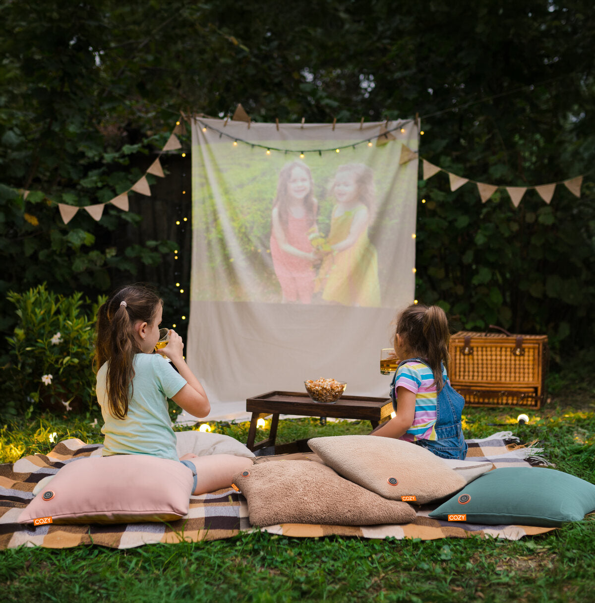 Two young girls sit outdoors on COZY Heated Cushion Cordless - UNA Velvet (45cm x 45cm) as they watch a projected movie at dusk. Snacks are on a small table, surrounded by string lights and bunting for a cozy atmosphere.