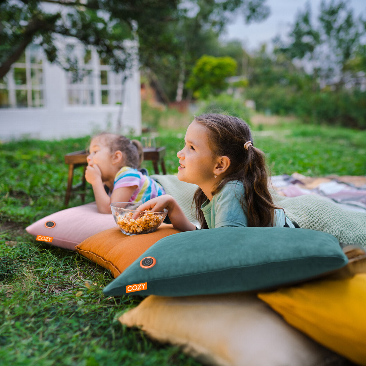 Two young girls relax outdoors on colorful pillows and the COZY Heated Cushion Cordless - UNA Velvet (45cm x 45cm), enjoying popcorn on a grassy lawn with trees and a white building in the background for a cozy atmosphere.