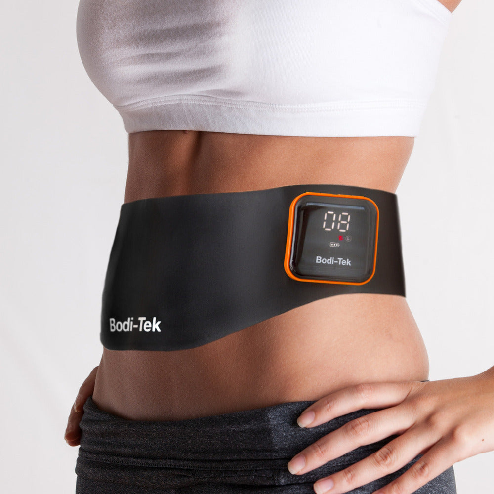 A person wears the Ab & Back Muscle Strengthener with heat, featuring a black design and digital display, over their bare midsection, styled with a white crop top and gray pants.